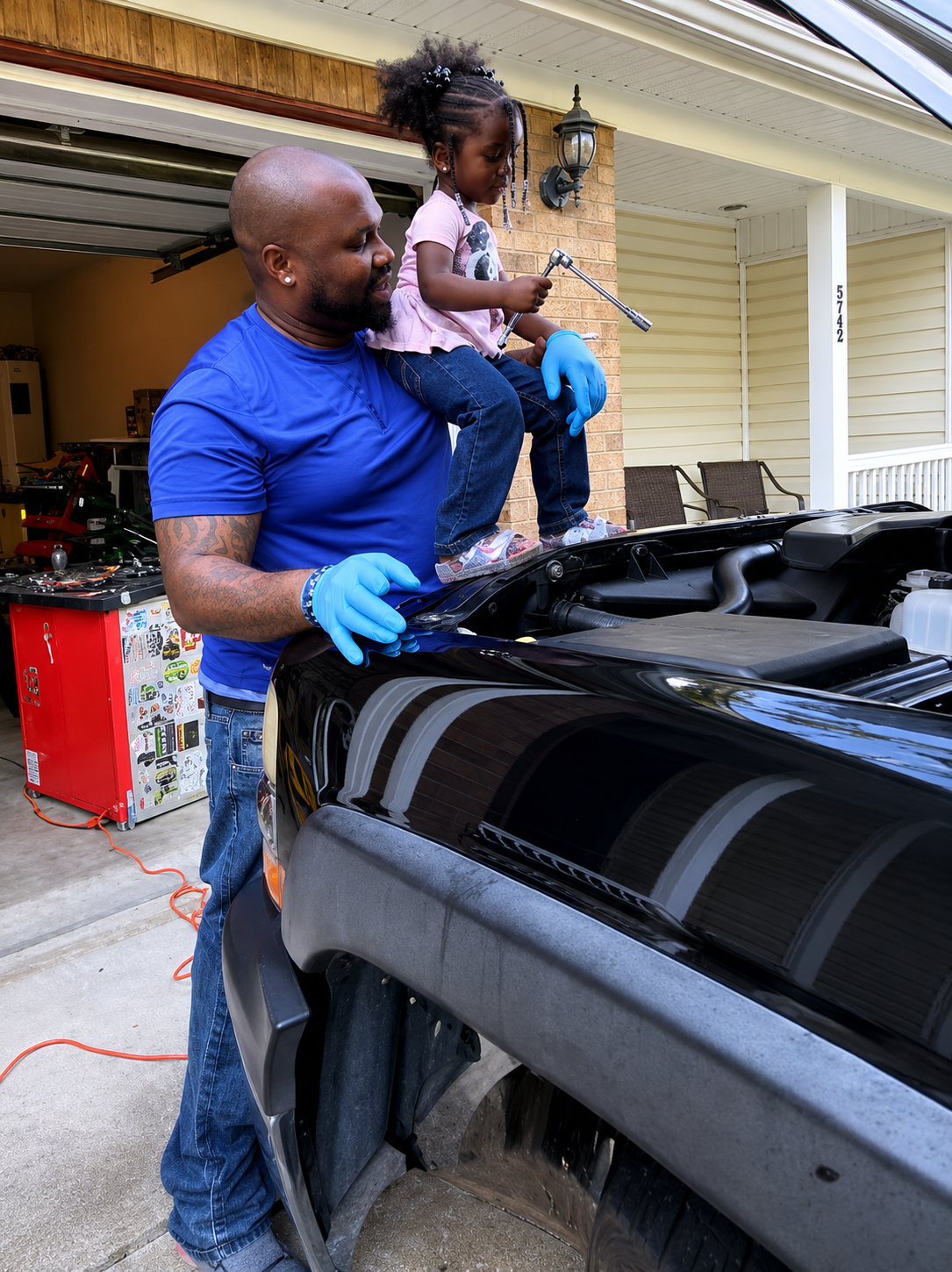 Man in blue shirt holding child while working on black vehicle in garage
